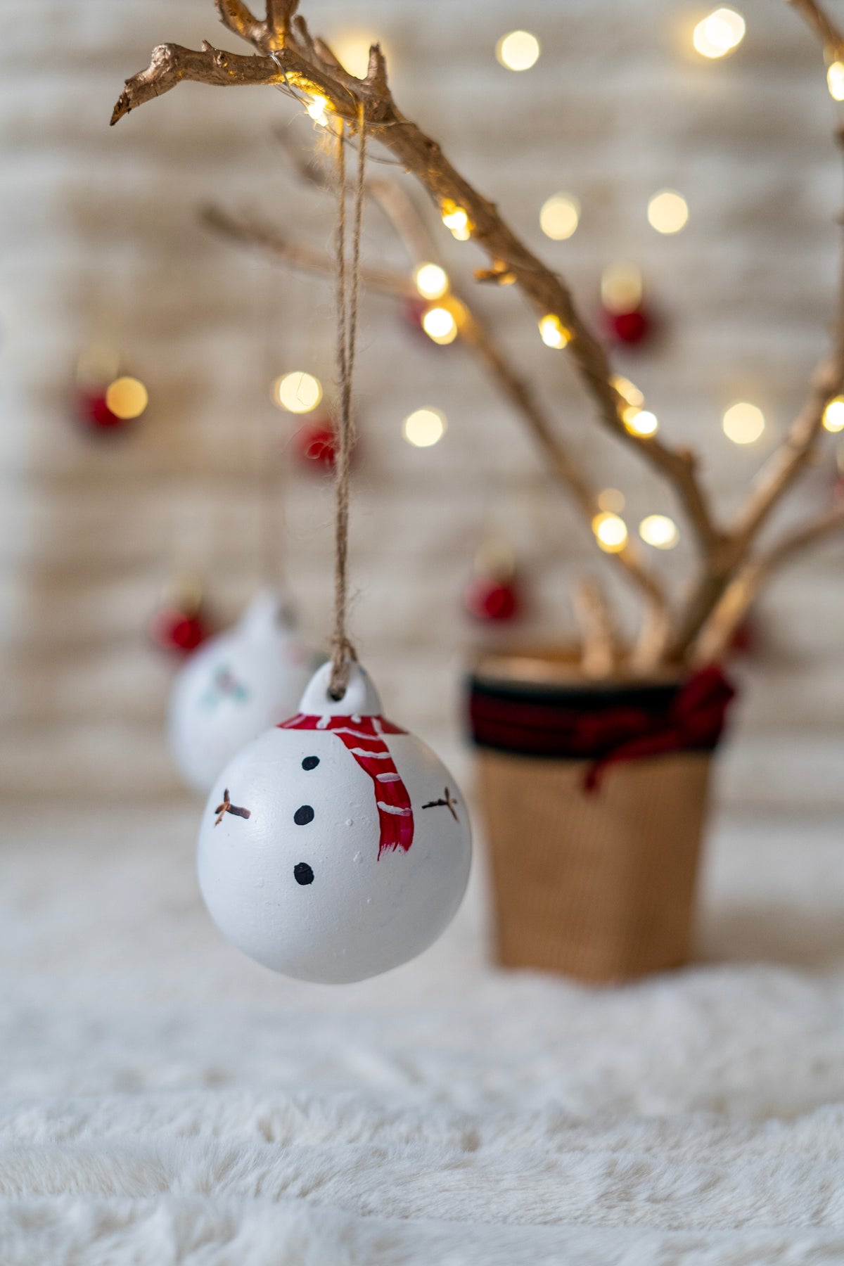 White snowman ornament hanging from a branch with blurred lights in the background